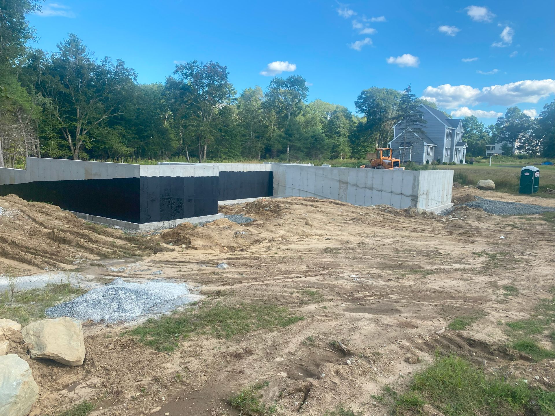 Construction site with concrete foundation walls, dirt lot, and trees under a blue sky