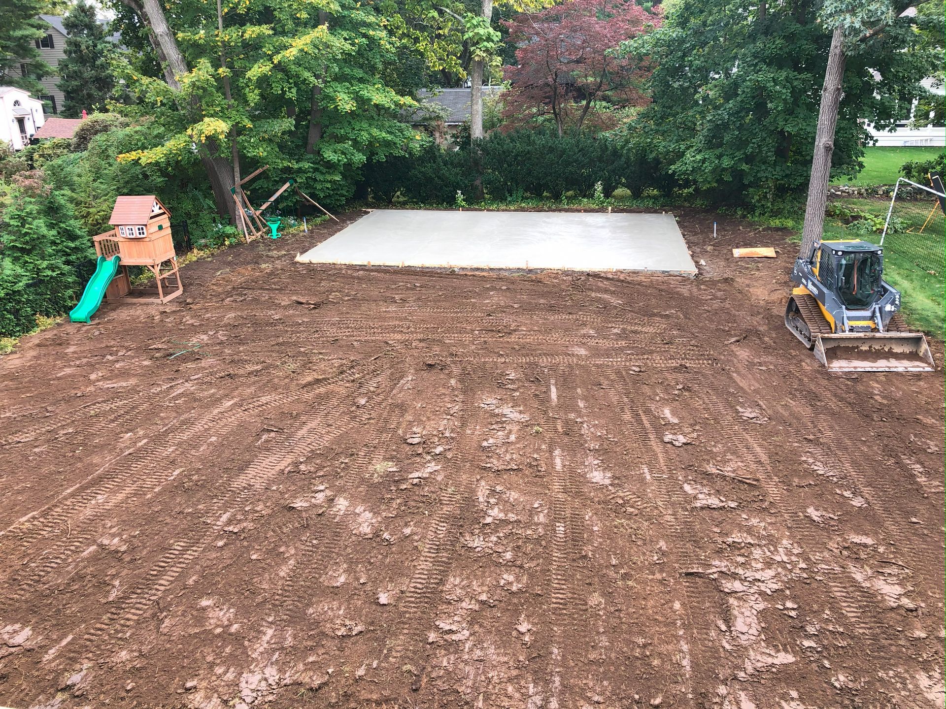 Backyard construction site with a dirt lot, concrete slab, and a small excavator near trees and shrubs