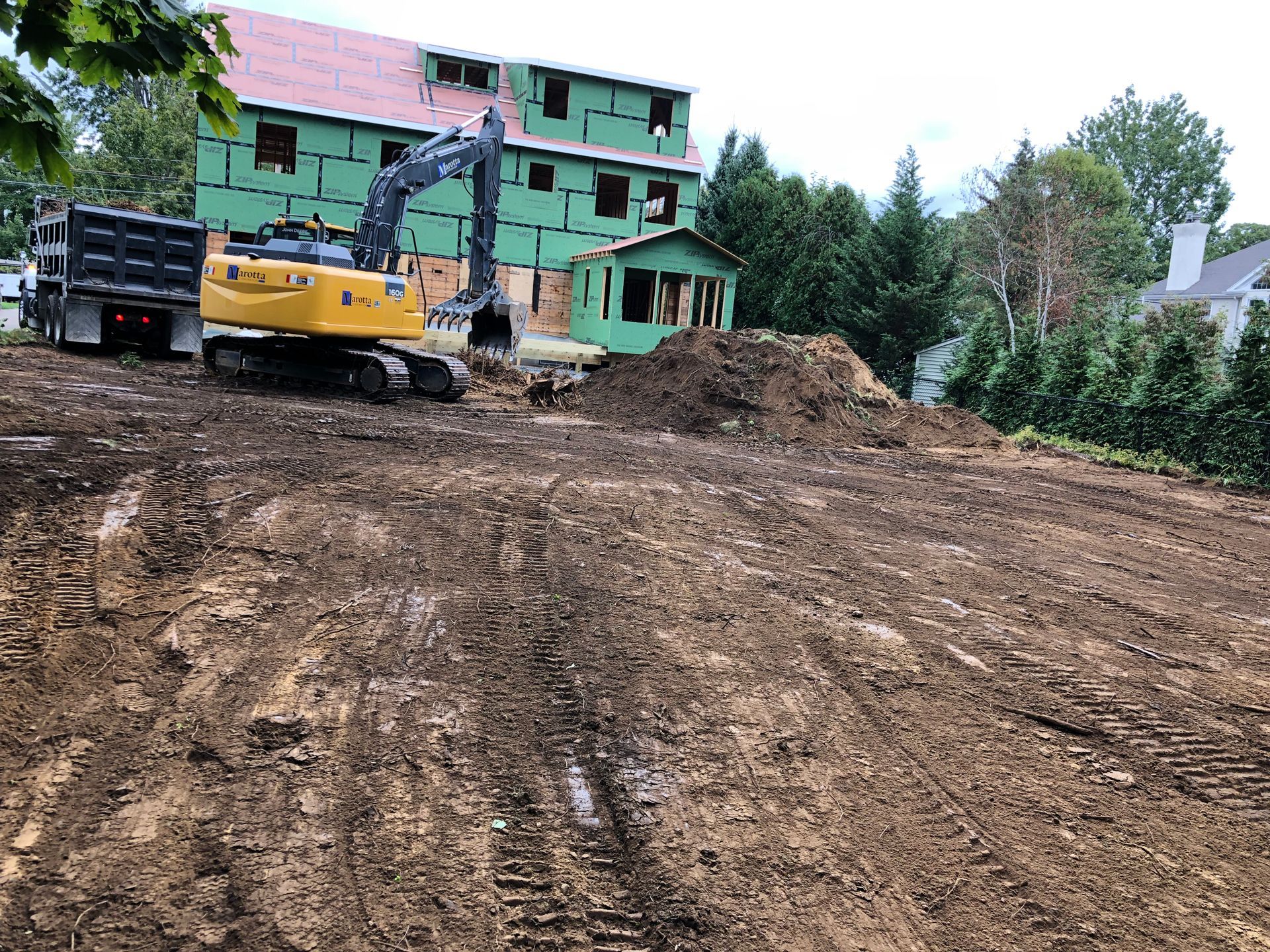 Construction site with excavator and truck on muddy ground beside a green building