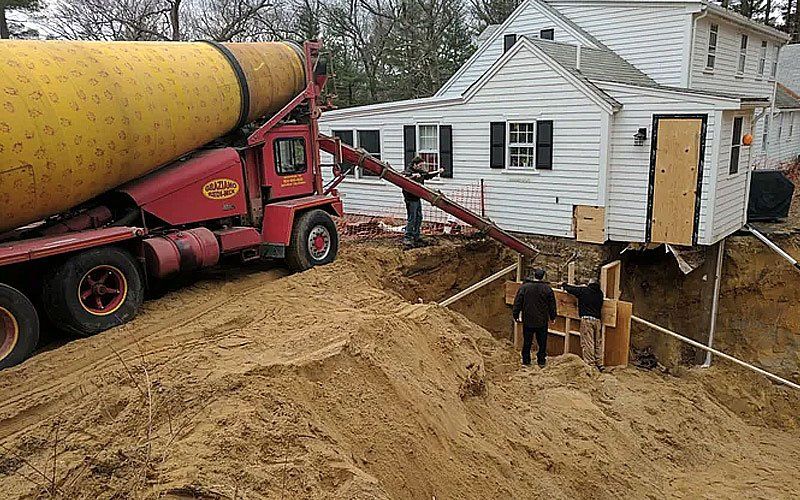 Large yellow concrete pump truck pours a hose into a house foundation beside a white home under construction