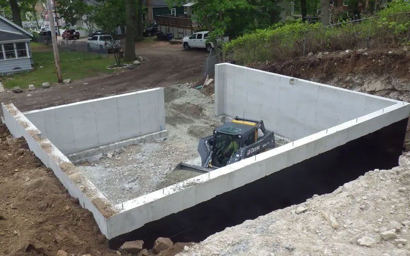 Concrete foundation walls at a construction site with a skid steer inside, surrounded by dirt and trees.