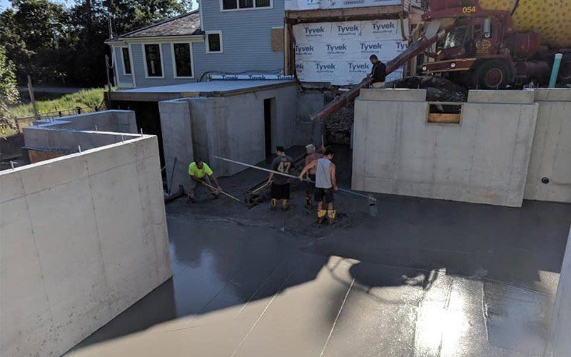 Workers pouring a fresh concrete slab in a residential construction site with retaining walls and a house in back