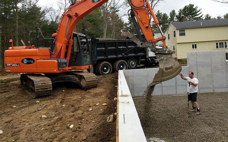 Orange excavator dumping gravel into a foundation trench beside a worker at a construction site