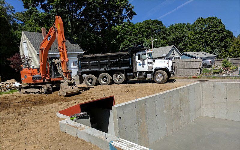 Excavator and dump truck at a construction site beside a large concrete pit and house foundations