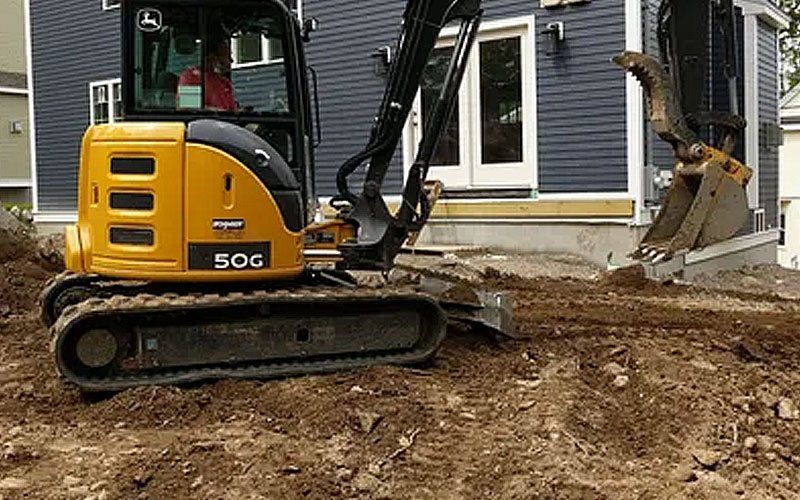 Yellow excavator beside a house, digging dirt near a foundation wall and window.