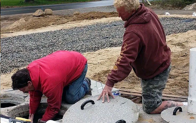 Two workers in hoodies kneel by concrete vault covers at a construction site.