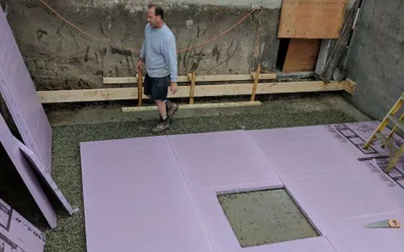 Man standing in a construction pit beside purple insulation panels and a square cutout in the floor