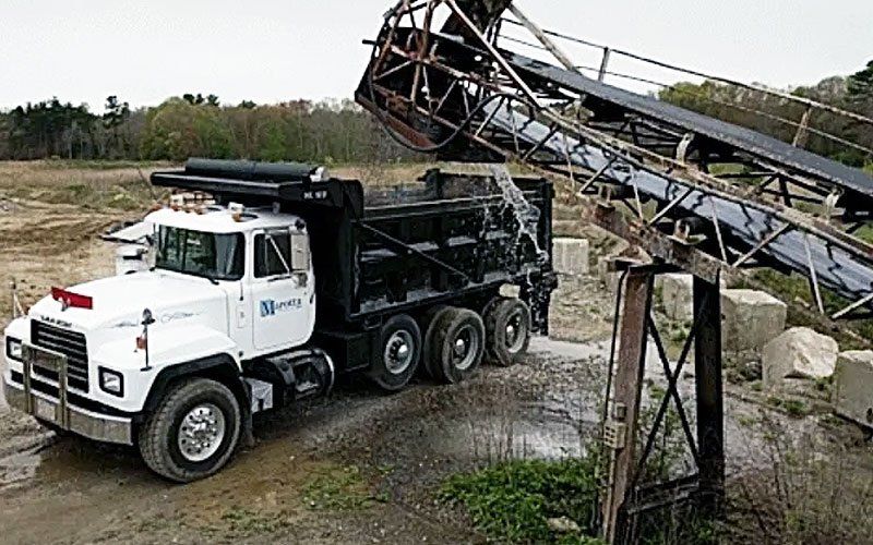 White dump truck beneath collapsed metal conveyor structure in a dusty industrial yard