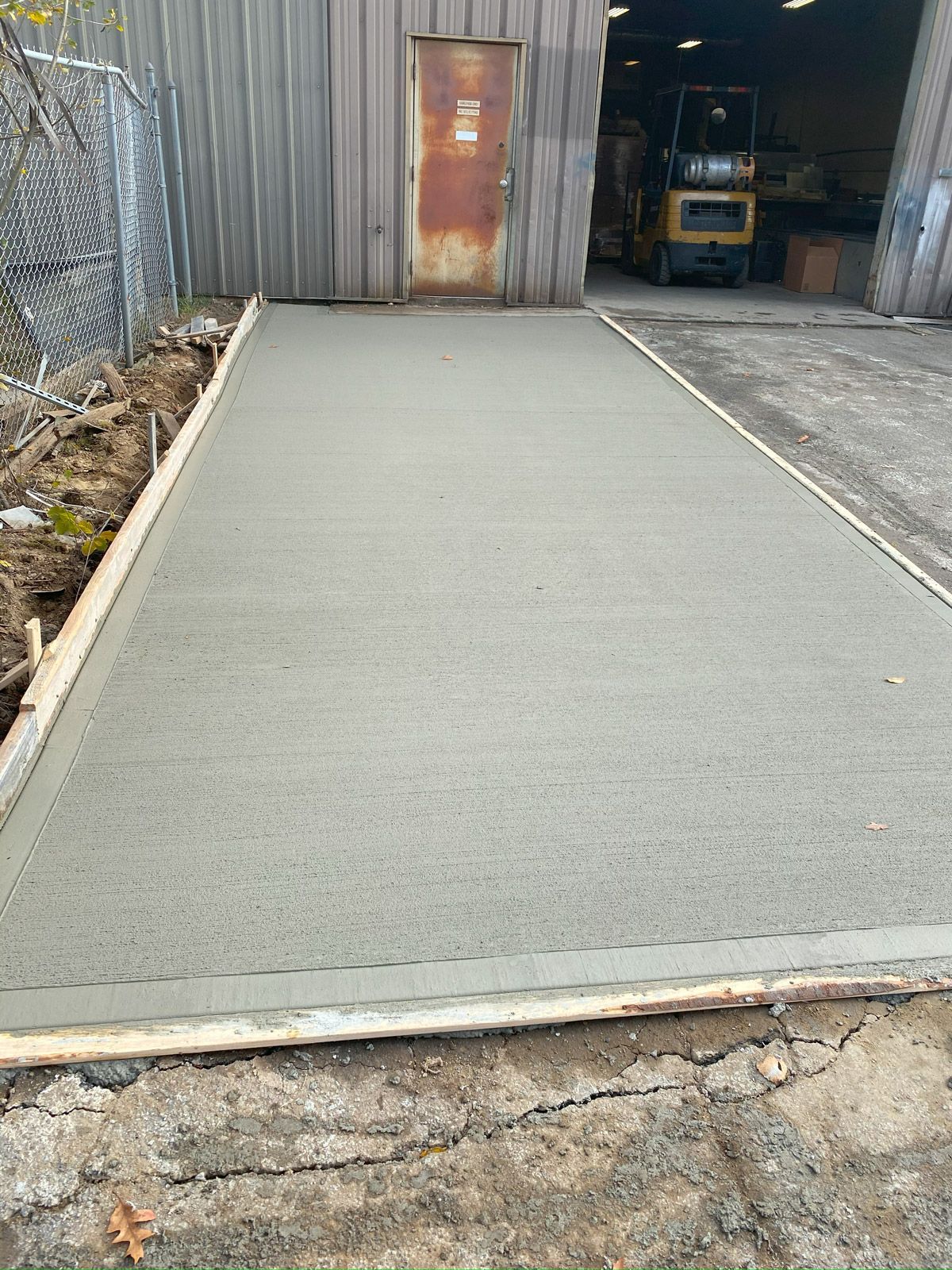Freshly poured concrete ramp leading to a warehouse entrance, with chain-link fence and equipment nearby.