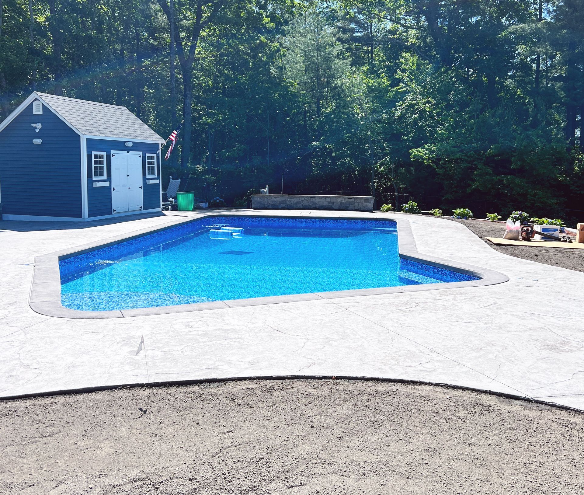 Backyard swimming pool with blue water, white patio, and a small blue pool house surrounded by trees
