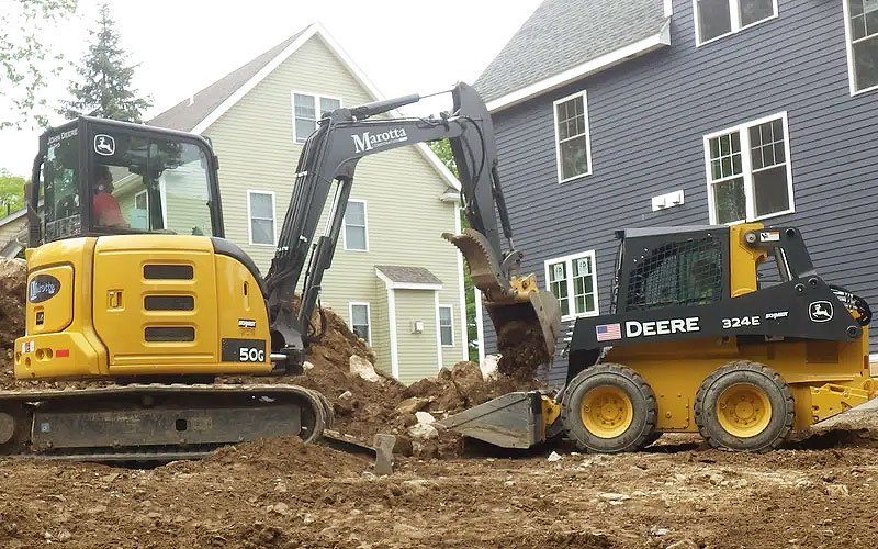 Two construction machines clearing dirt beside a house, with a yellow excavator and black skid steer.