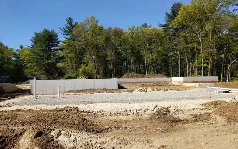 Construction site with concrete foundation walls and dirt lot bordered by trees under a clear sky