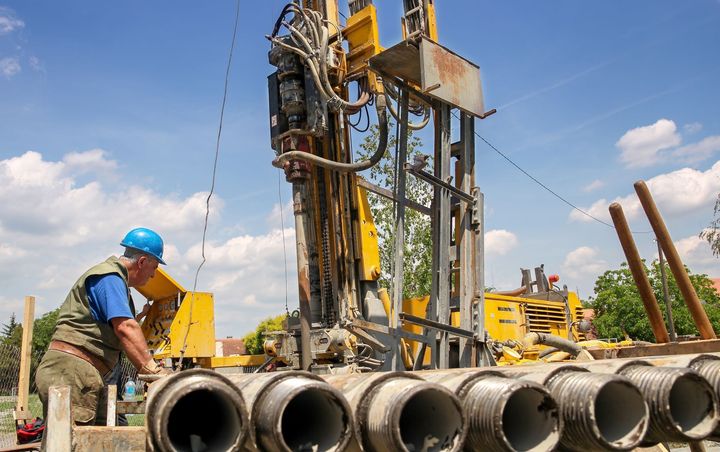 Worker and drilling rig at a construction site with stacked concrete pipes under a blue sky