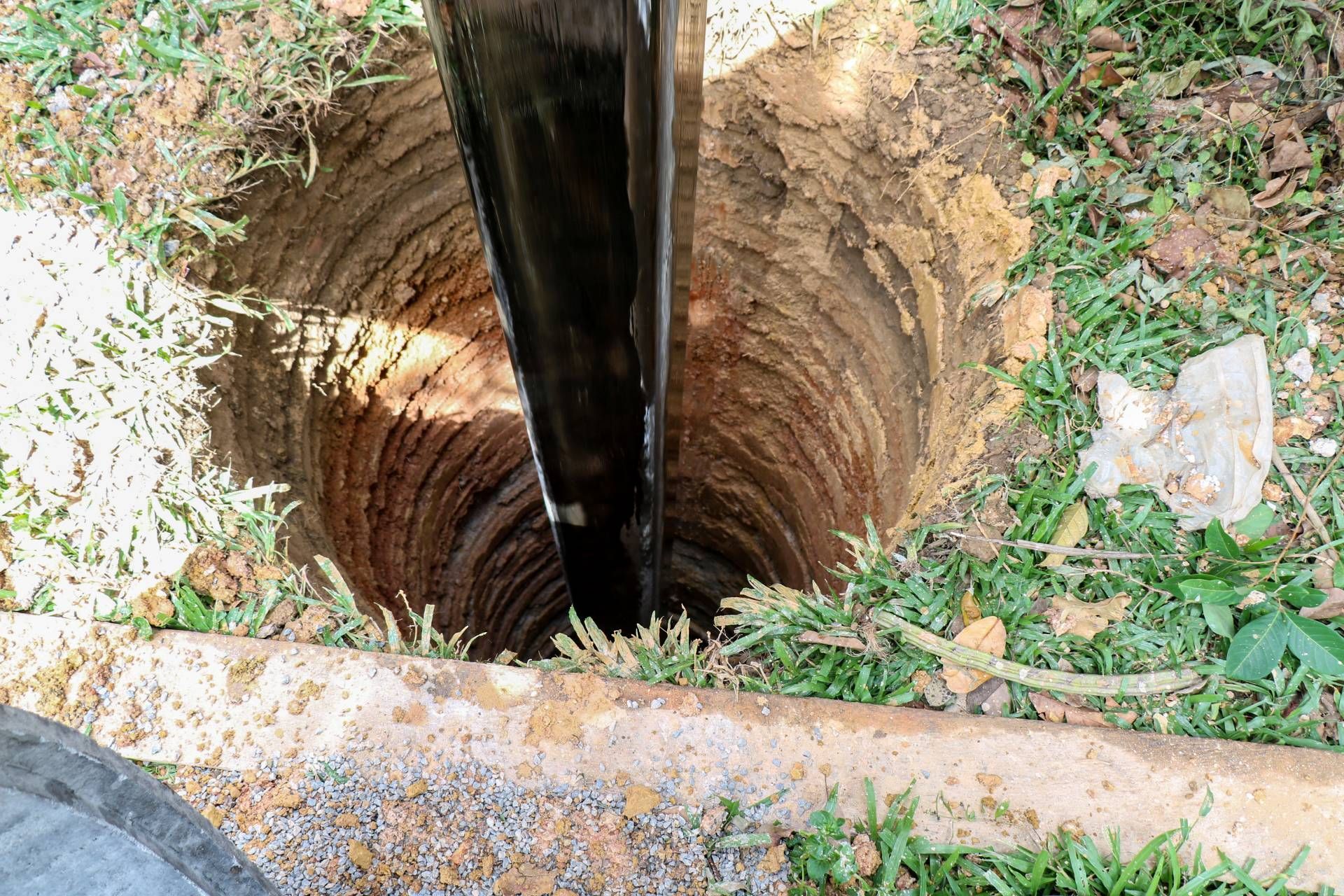 Deep narrow well or shaft opening with brick walls and a dark pipe in the center, surrounded by grass and stone.