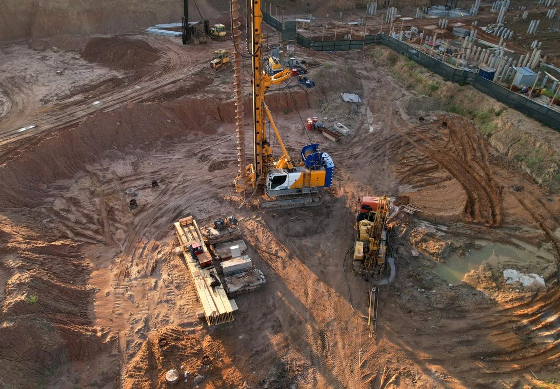 Construction site with drilling rig, excavator, and workers on muddy earthworks area