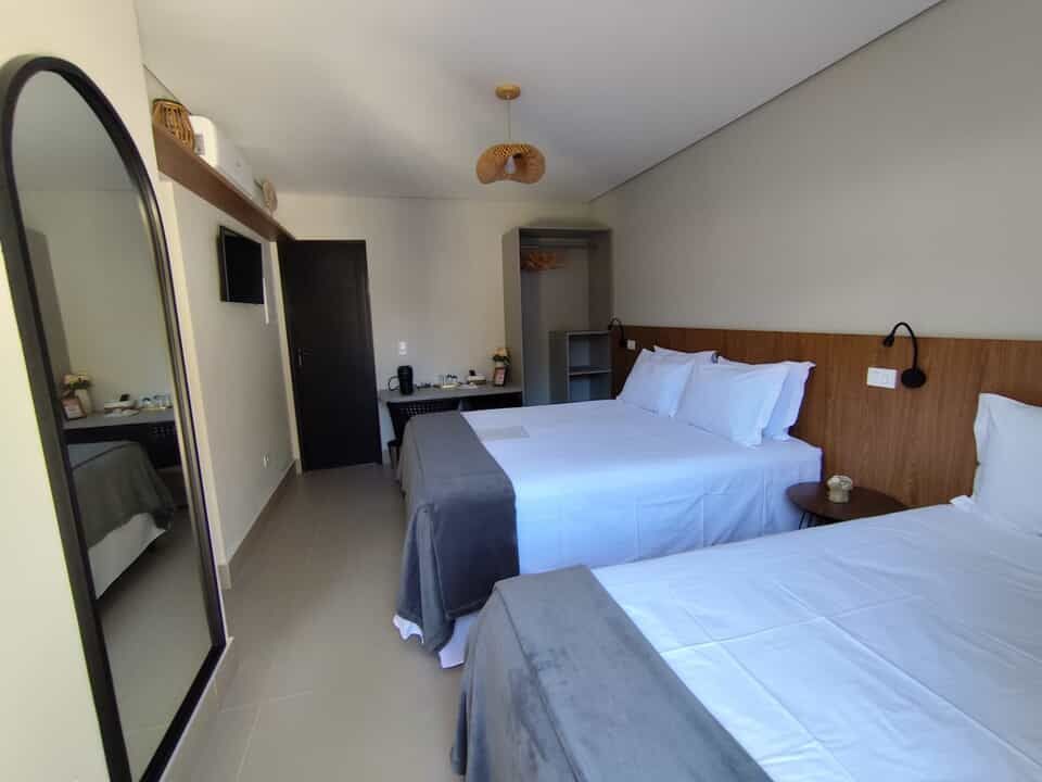 Quarto de hotel moderno com duas camas arrumadas, escrivaninha, espelho e TV de parede.