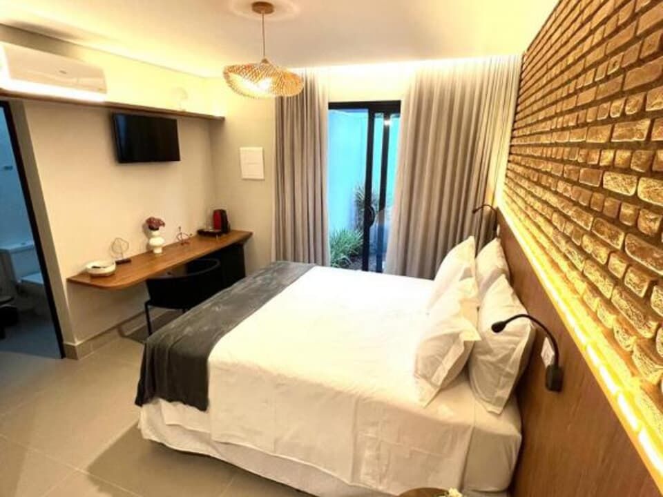 Quarto de hotel moderno com parede de tijolos à vista, cama arrumada, escrivaninha, TV e portas para varanda.
