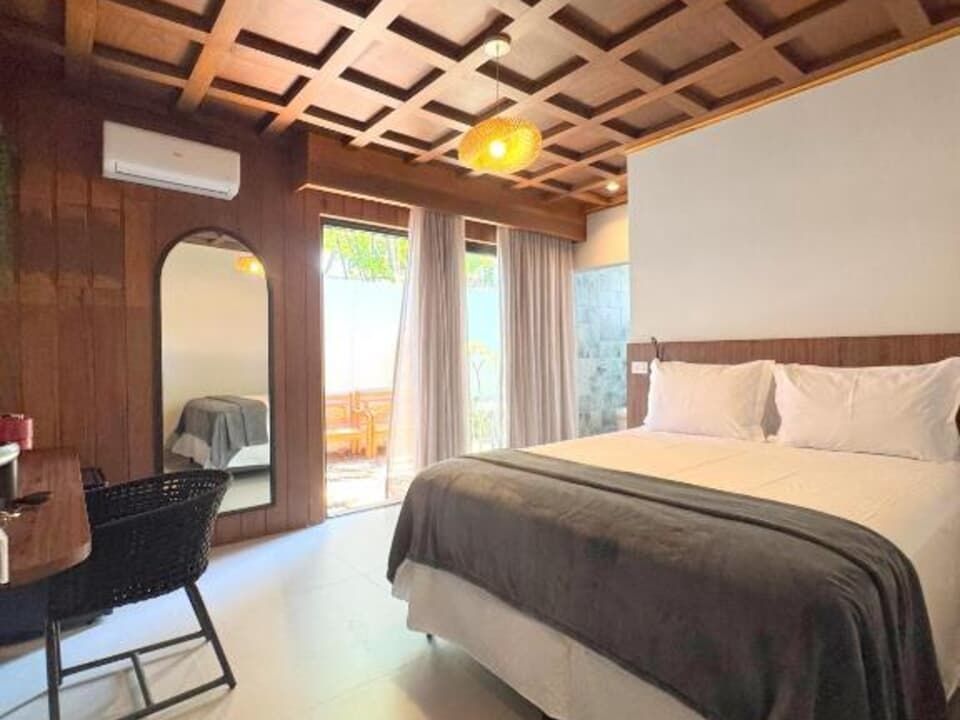 Quarto luminoso com uma cama grande, teto de madeira, escrivaninha, espelho e portas de correr que dão para uma varanda.