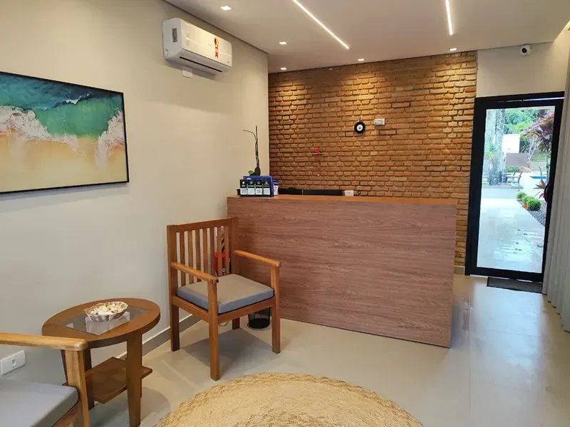 Pequena área de recepção com uma mesa de madeira, cadeira, mesa lateral, obras de arte na parede e uma porta de vidro.