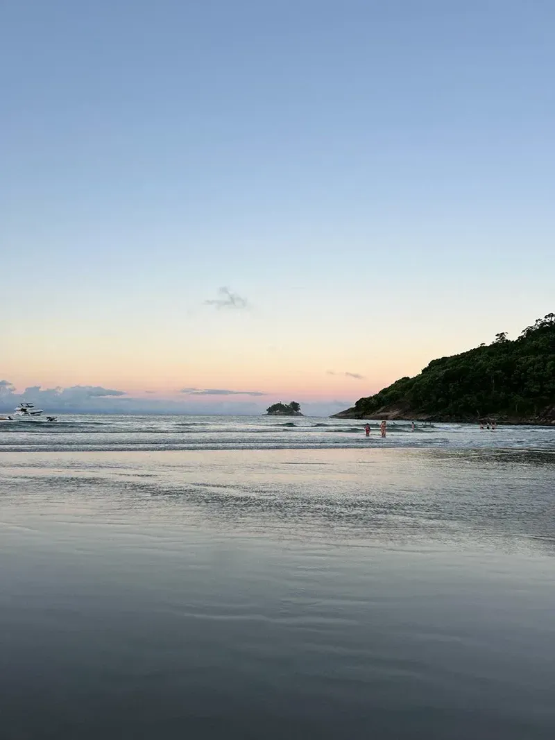 Praia tranquila ao entardecer, com céu em tons pastel, ondas suaves e uma orla arborizada.