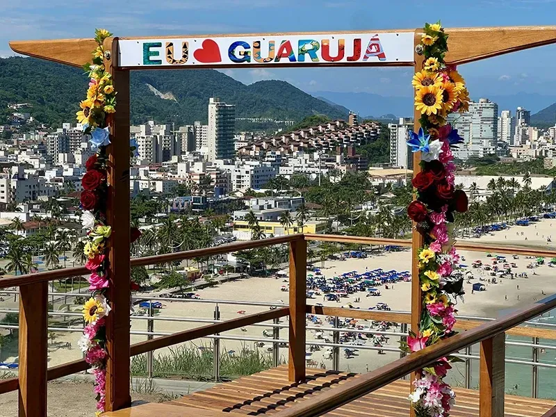 A moldura colorida com a inscrição “EU AMO GUARUJÁ” oferece uma vista panorâmica da praia, do horizonte da cidade e das montanhas verdejantes.