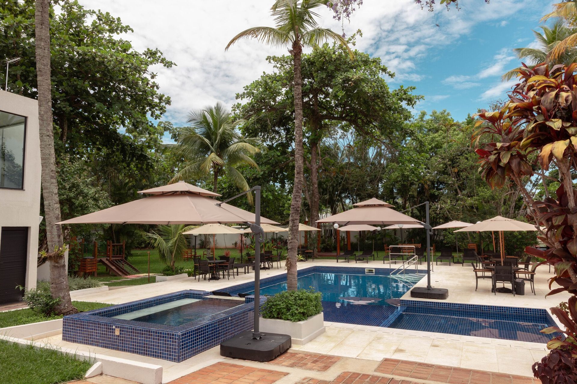 Piscina do resort com azulejos azuis, palmeiras e espreguiçadeiras bege sob guarda-sóis.