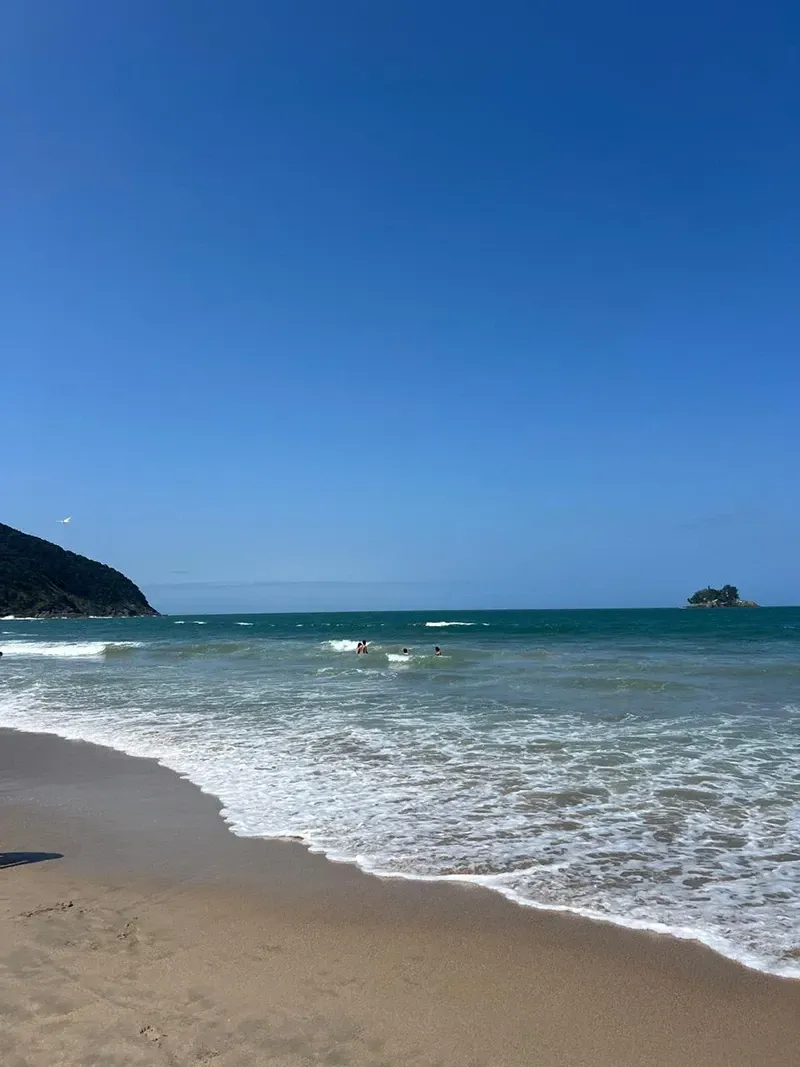 Praia ensolarada com ondas suaves, céu azul e uma pequena ilha no horizonte.