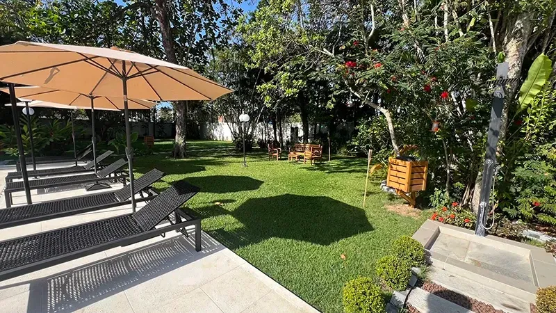 Pátio com espreguiçadeiras, guarda-sol bege e um jardim verdejante sob um sol radiante.