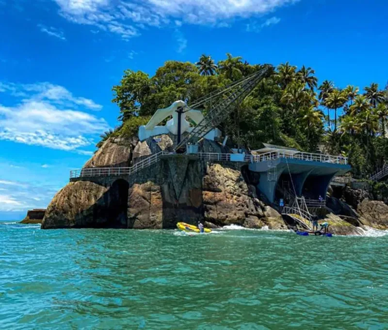 Ilha rochosa costeira com uma ponte de concreto, palmeiras exuberantes e oceano azul sob um céu claro.