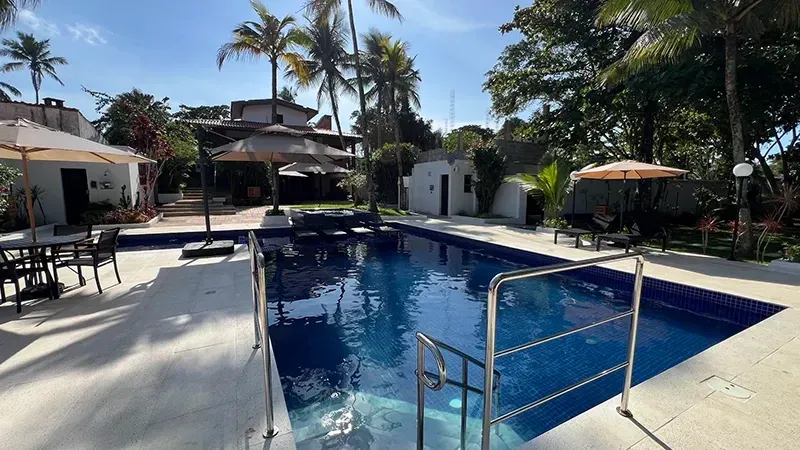 Piscina ensolarada de resort com água azul, espreguiçadeiras, palmeiras e cabanas brancas.