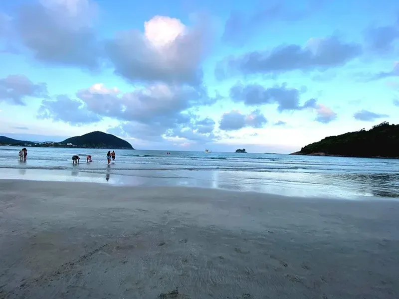 Pessoas caminhando em uma praia tranquila ao entardecer sob um céu azul e nublado.