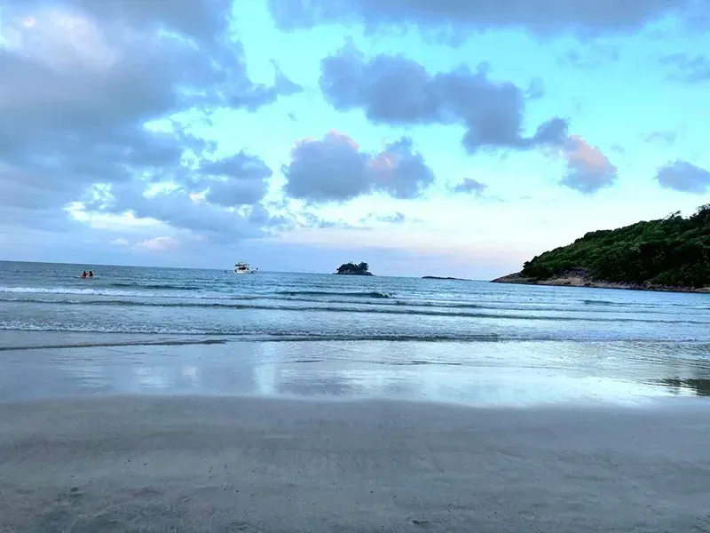 Praia tranquila ao entardecer, com ondas suaves, céu azul nublado e uma pequena ilha rochosa no horizonte.