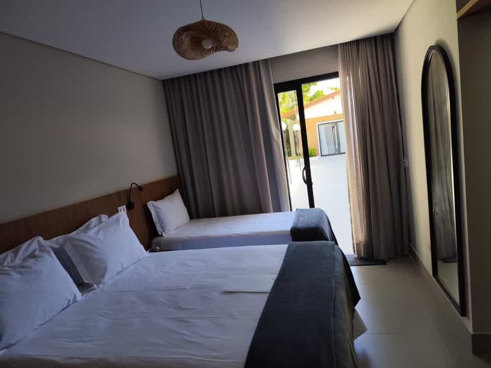 Quarto de hotel com duas camas bem arrumadas, cortinas cinzentas e uma porta de varanda aberta que permite a entrada de luz natural.