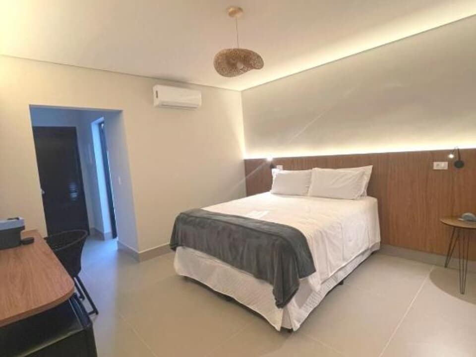 Quarto de hotel moderno com cama arrumada, parede com detalhes em madeira, escrivaninha e iluminação aconchegante no teto.