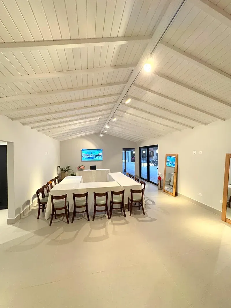 Sala de jantar iluminada com mesa comprida, cadeiras, teto branco alto e TV fixada na parede.