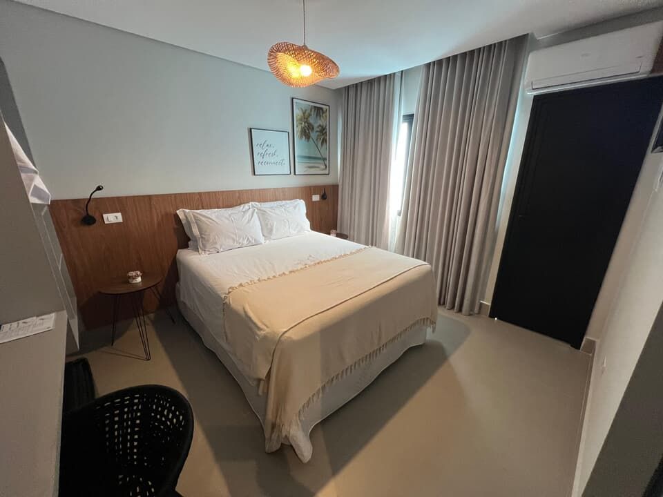 Quarto de hotel moderno com cama arrumada, cortinas, mesa de cabeceira e luminária pendente aconchegante.