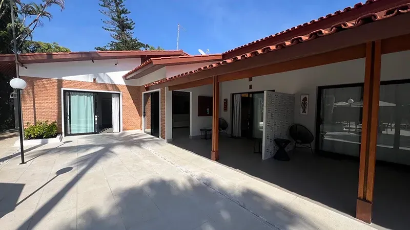 Pátio ensolarado com casa de telhado vermelho, paredes brancas e área de estar externa sombreada.