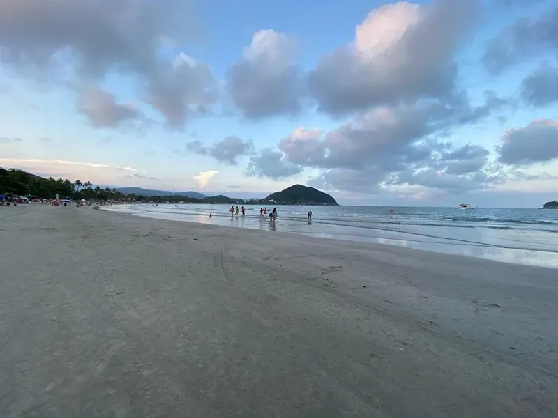 Ampla praia de areia ao pôr do sol, com pessoas dispersas, ondas calmas e uma pequena ilha no horizonte.