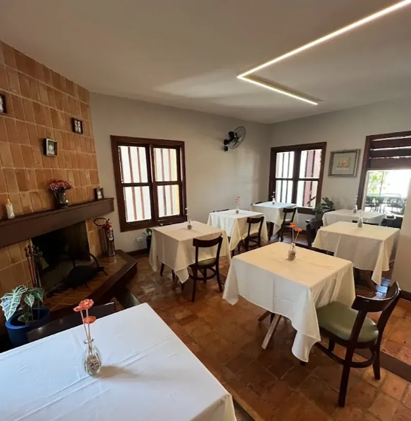 Sala de jantar aconchegante com mesas cobertas com toalhas brancas, cadeiras de madeira, lareira e iluminação suave no teto.