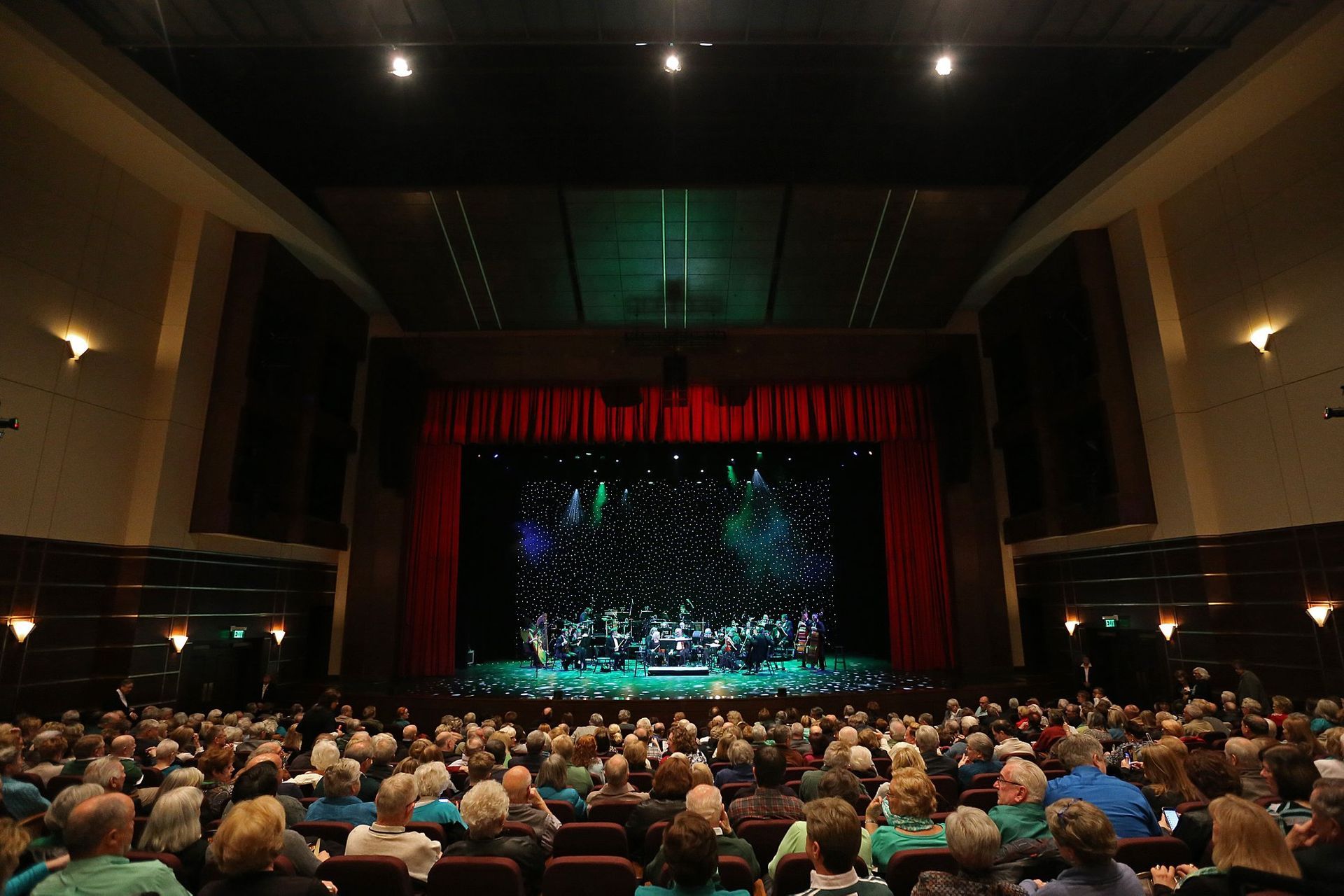Auditorium AV solutions. Audience watches a stage show in a large theater, red curtain, dark walls, overhead lights.