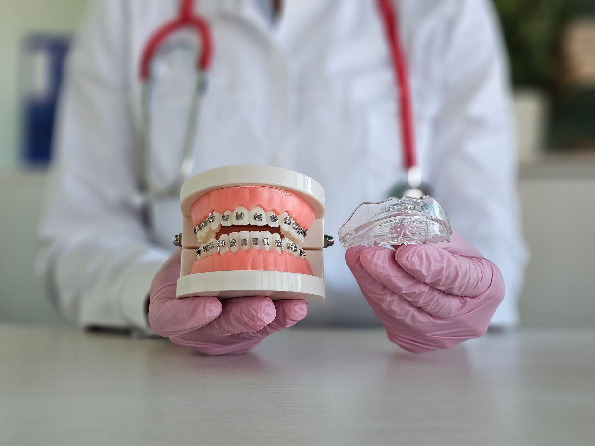 Dentist holding a model with braces and an Invisalign retainer. Pink gloves, red stethoscope.