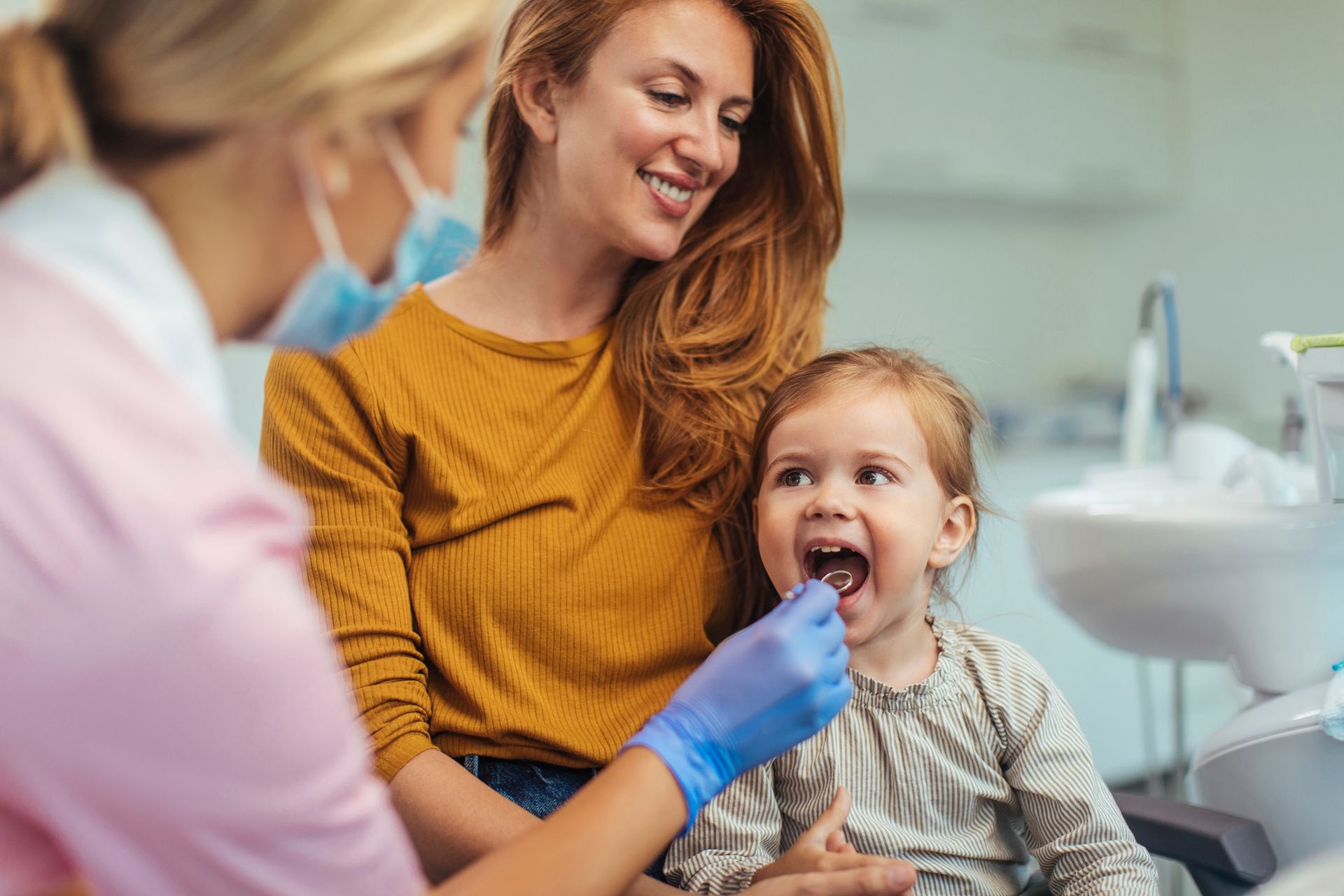 Dentist examining a child's teeth, mother watches. Dentist, light skin; mother, red hair; child smiles in the dental chair.
