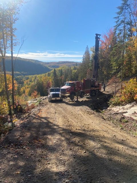 Installation de forage et camion d'assistance sur un chemin de terre dans une zone boisée offrant une vue pittoresque sur la vallée.