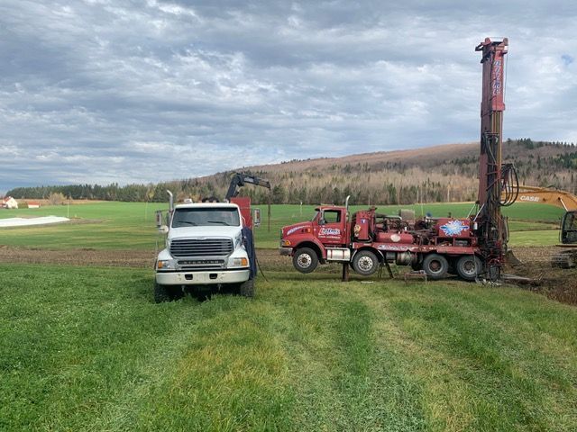 Plateforme de forage sur un champ verdoyant sous un ciel nuageux, à côté d'un camion blanc.