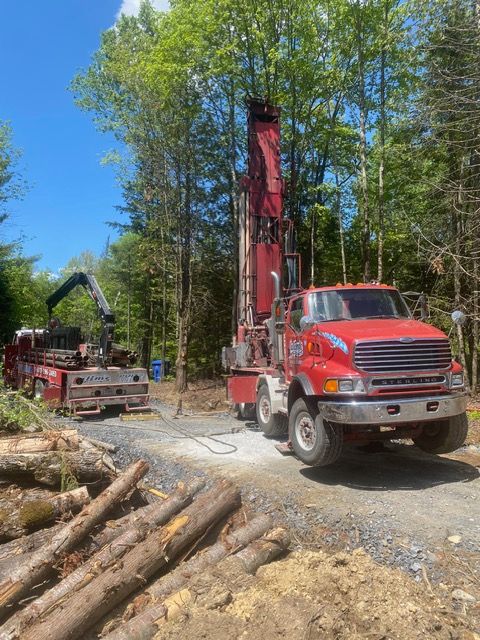 Un camion de forage rouge et un camion d'assistance sur un chemin de terre dans une zone boisée, en train de forer un puits.