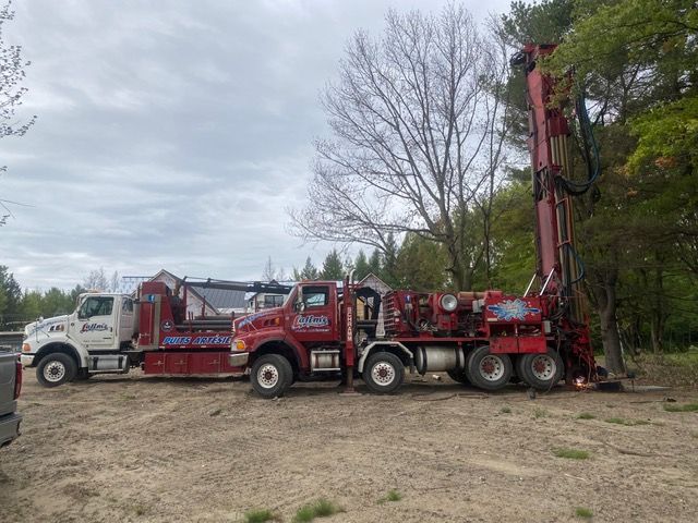 Camion de forage rouge et blanc à côté d'un arbre, en extérieur par temps nuageux.