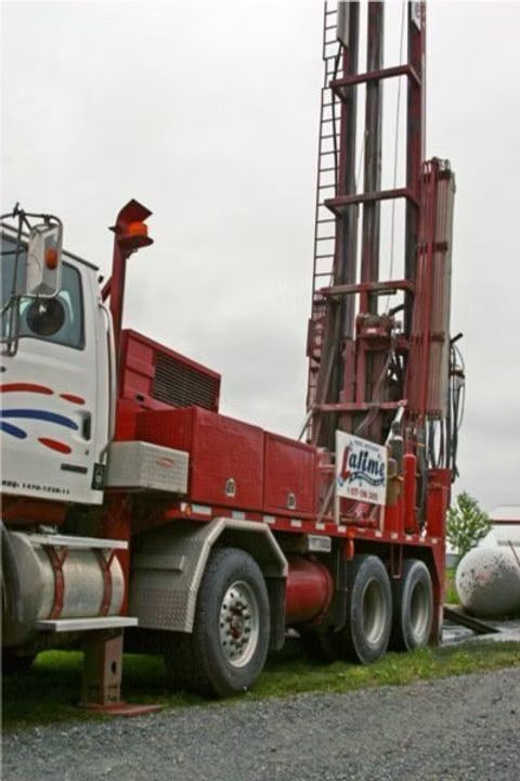 Une foreuse rouge et blanche montée sur un camion, avec une haute tour, dans un cadre rural.