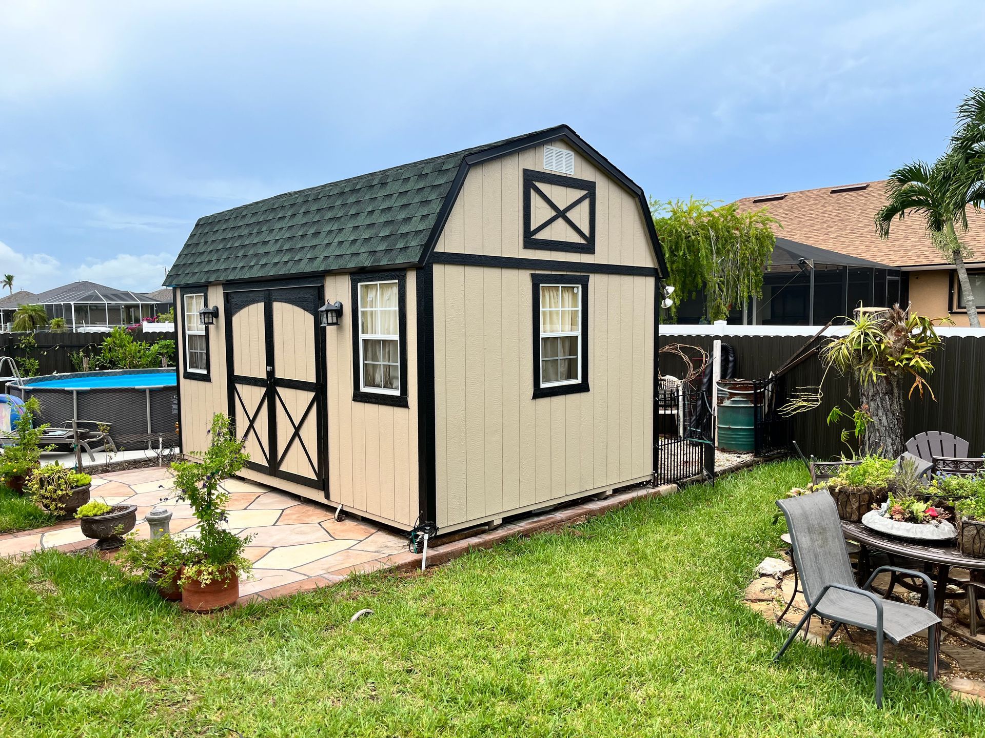 Tan shed with black trim and green roof in a backyard, near pool and fence.