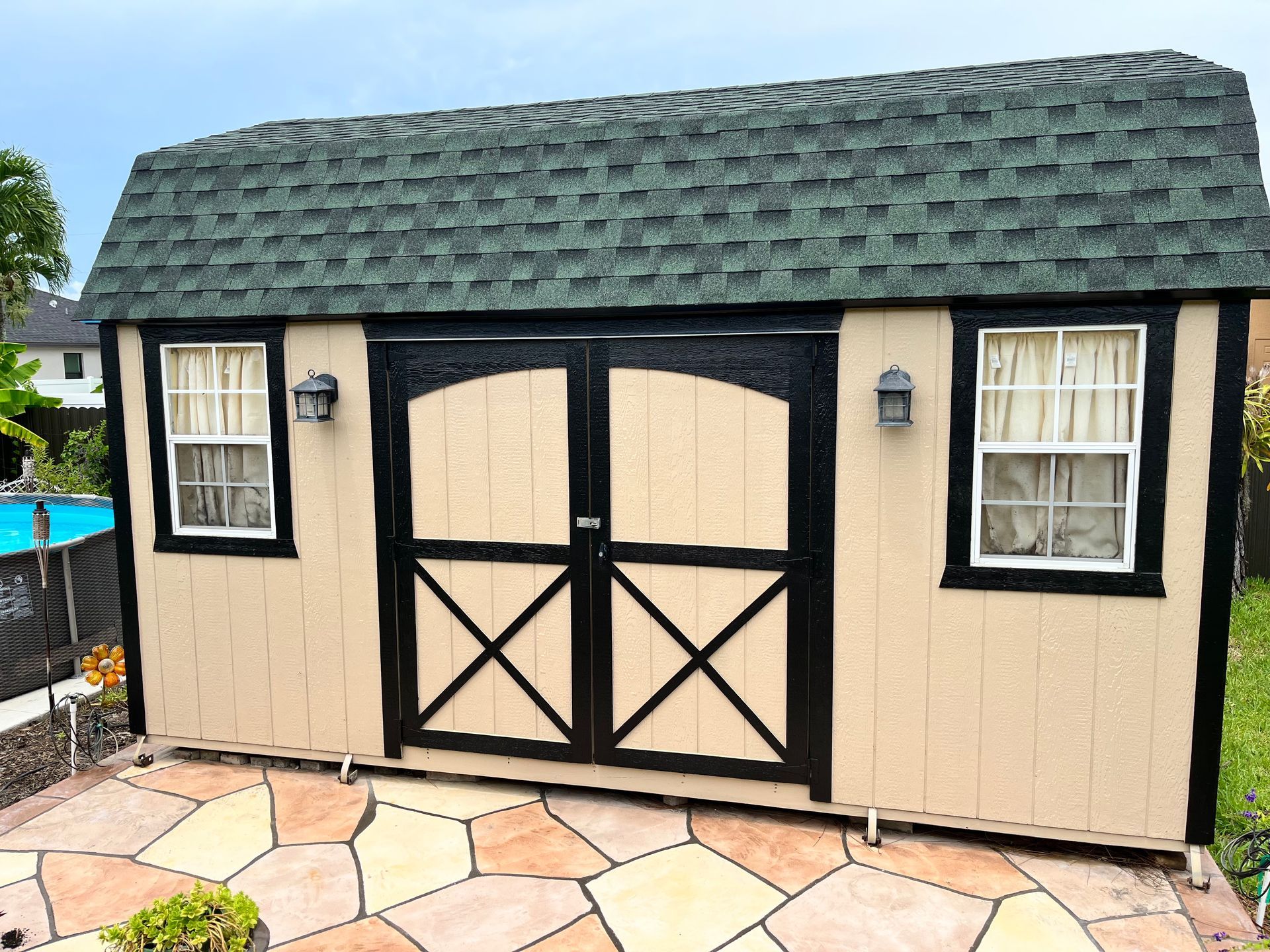 Tan shed with black trim and door, green roof, white-framed windows, set on a stone patio.