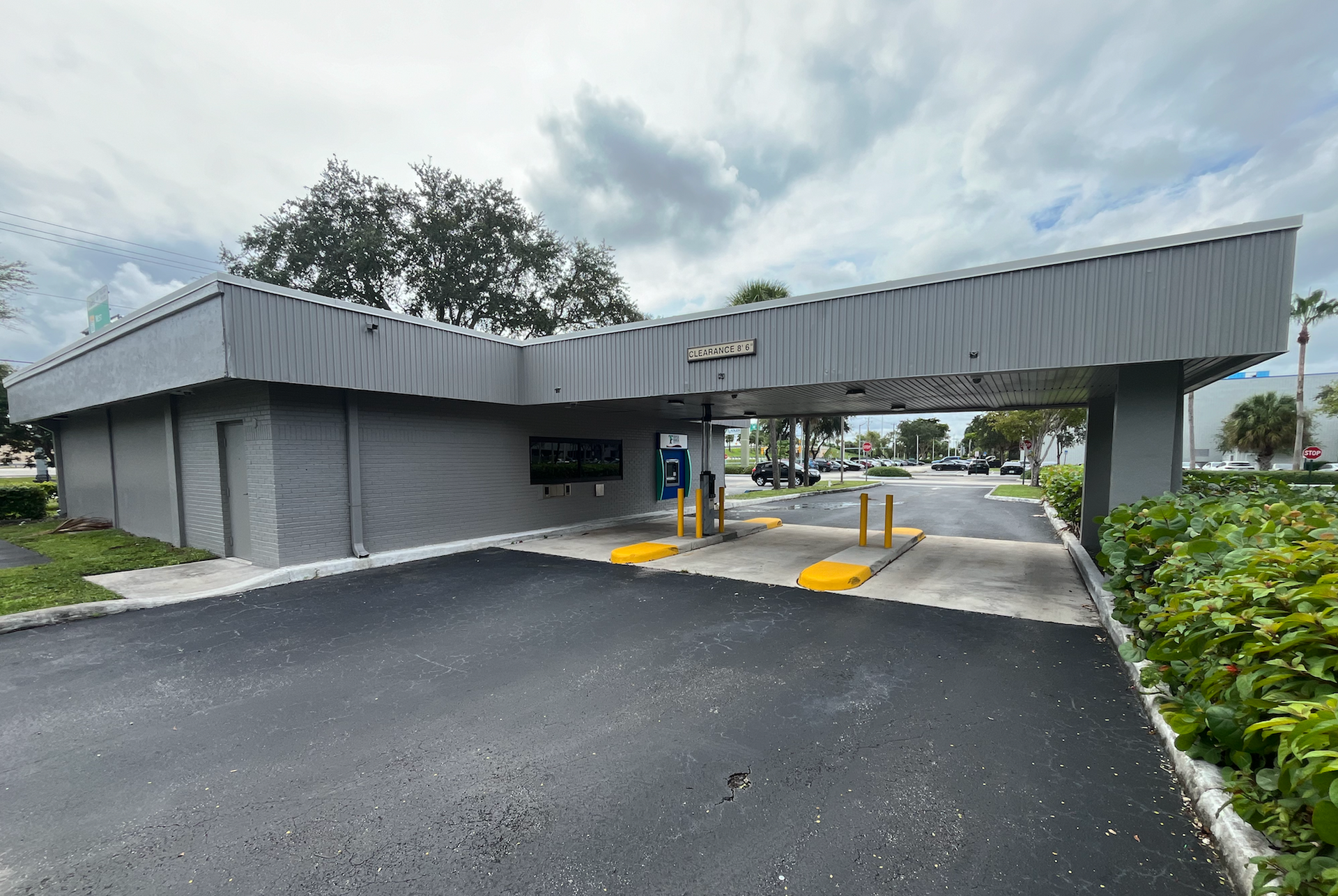 Drive-thru bank building with a gray facade under a cloudy sky. Asphalt driveway leads to the service window.